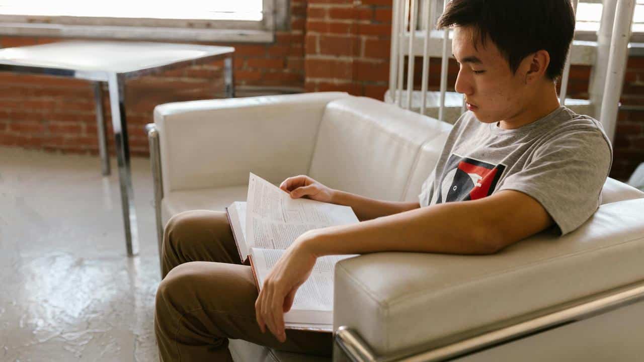 A young man sitting on a couch reading a book.