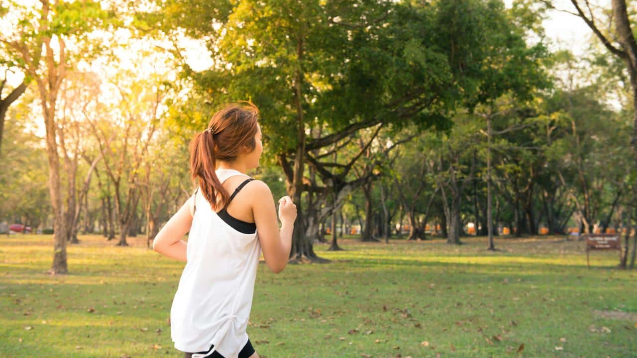 A woman jogging through a sunlit park with trees and grass.