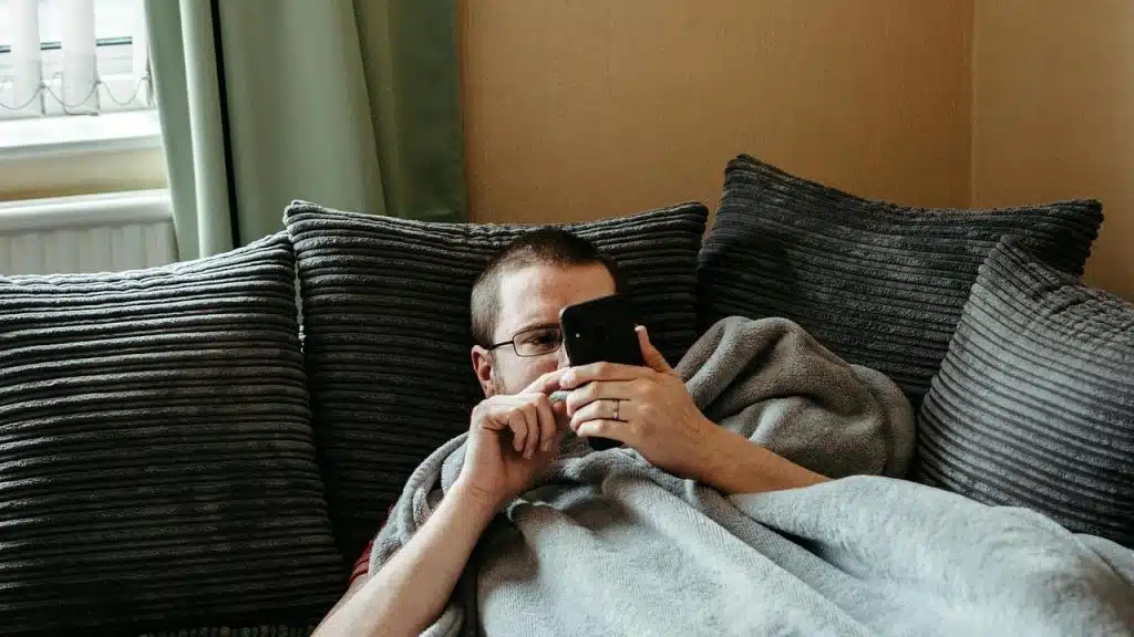A man lying on a couch under a blanket, looking at his phone.