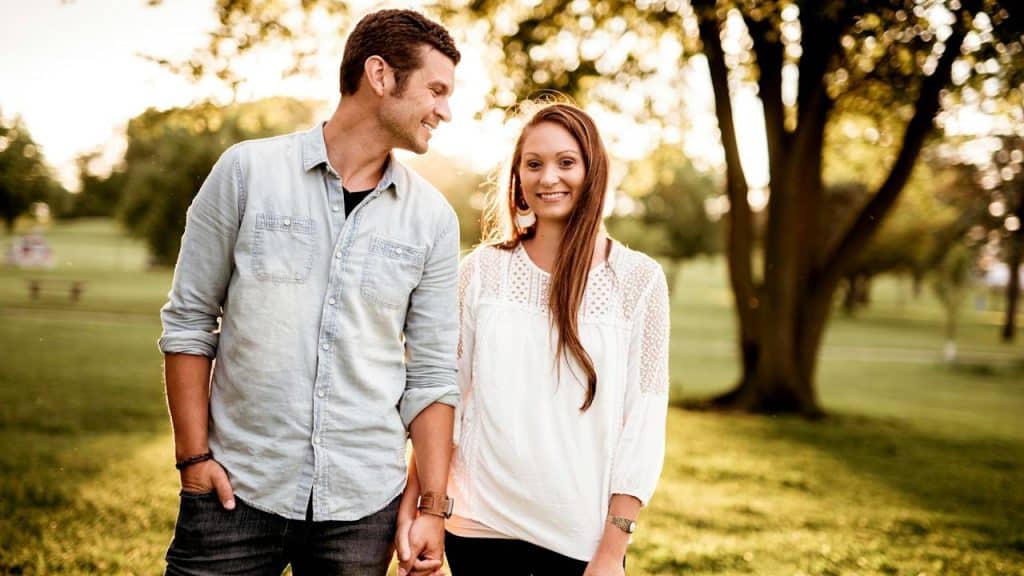 A man looking at a woman's hand and looking towards her while they are standing in a park.