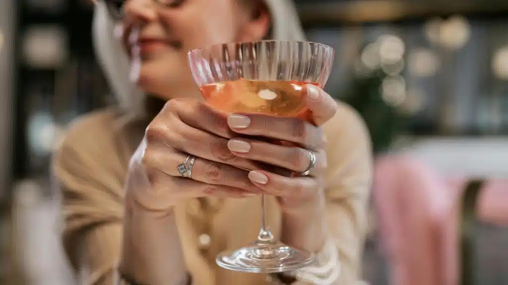 A person holding a cocktail glass with both hands, smiling in the background.