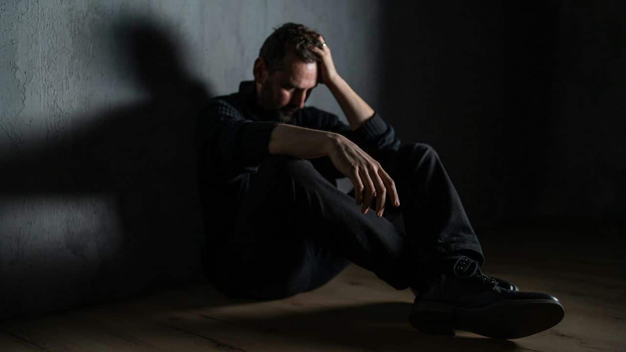 A man sits alone in a dark room with his hand on his head, looking stressed and sad