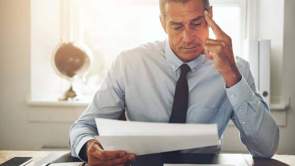 A man in a shirt and tie sits at a desk looking at papers with a hand to his temple.
