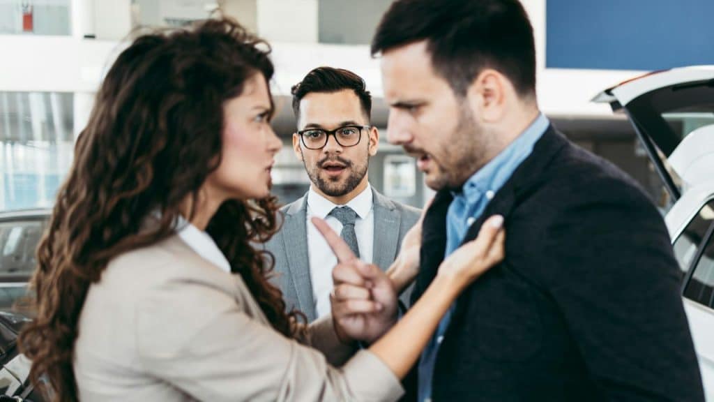 A woman gestures at a man’s jacket while another man in a suit looks on behind them.