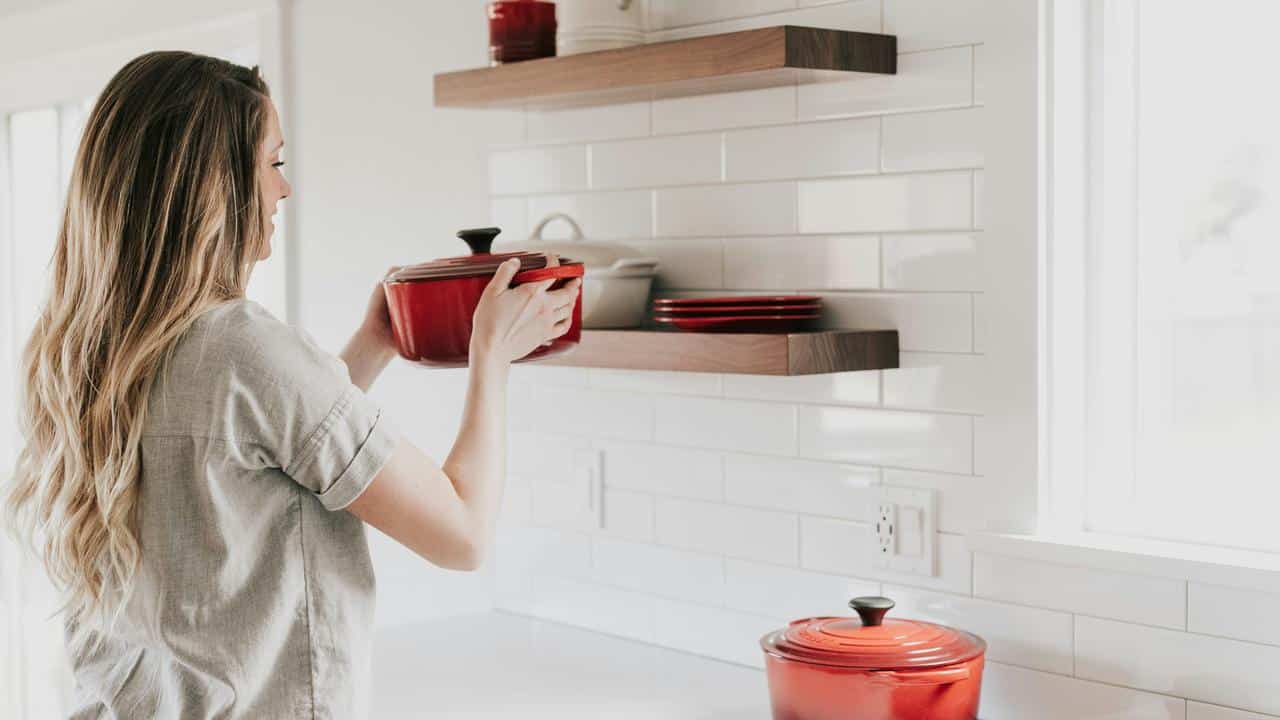 A woman placing a red pot on a kitchen shelf.