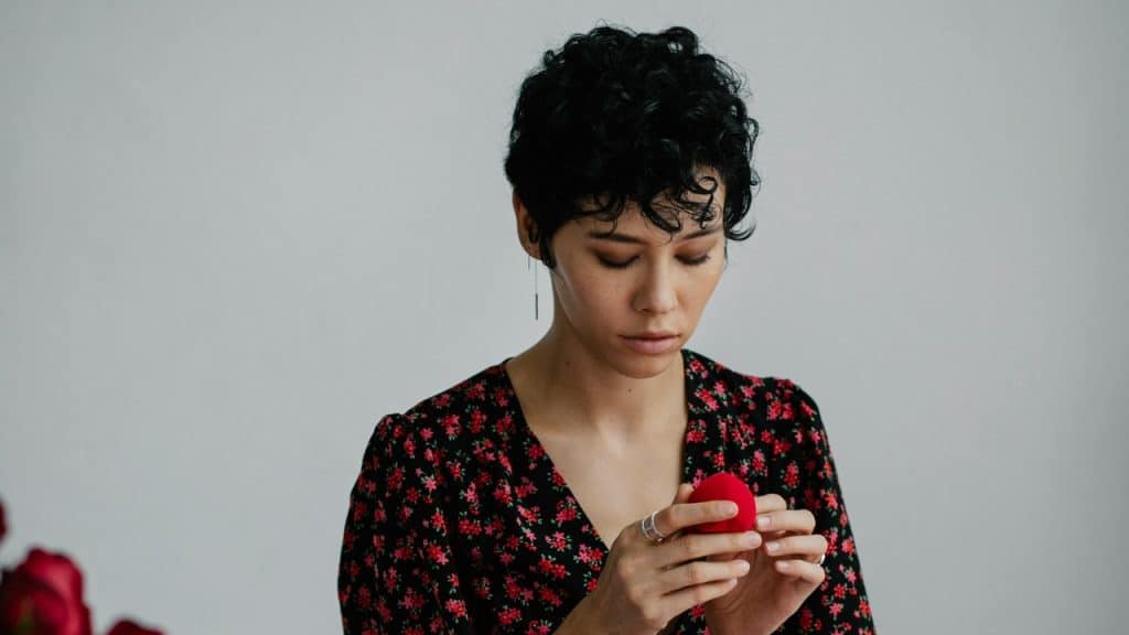 A woman with short curly hair looks down while holding a small red velvet box.