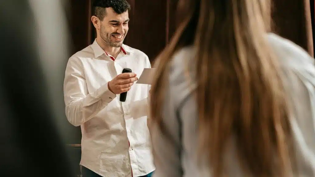 A smiling man holding a microphone and reading from a card.