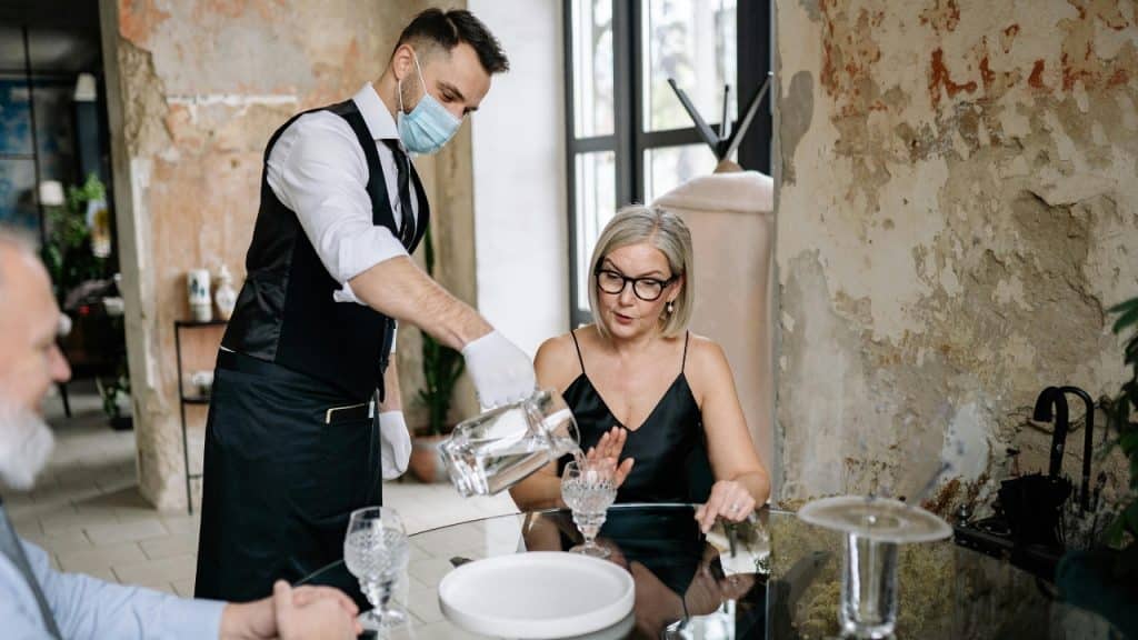A waiter wearing a mask pours water for a woman sitting at a restaurant table.