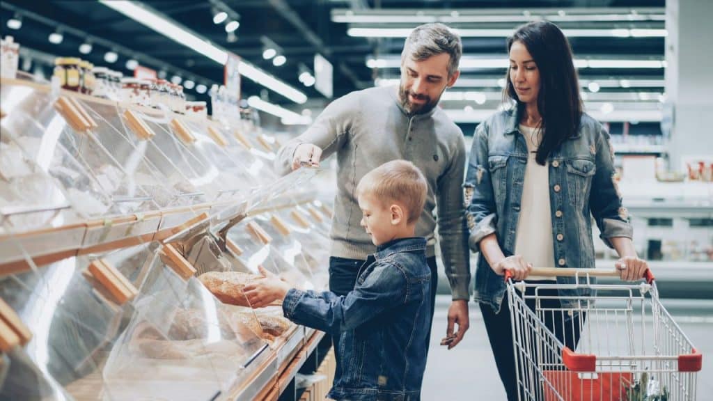 A man, woman, and young boy stand together in a grocery store selecting bread from bins.