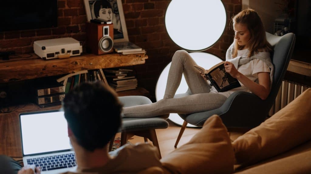 A woman reads a book in a chair while a man works on a laptop.