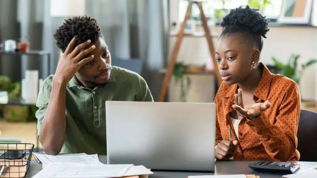 A man and woman sit at a desk looking at a laptop with focused expressions.