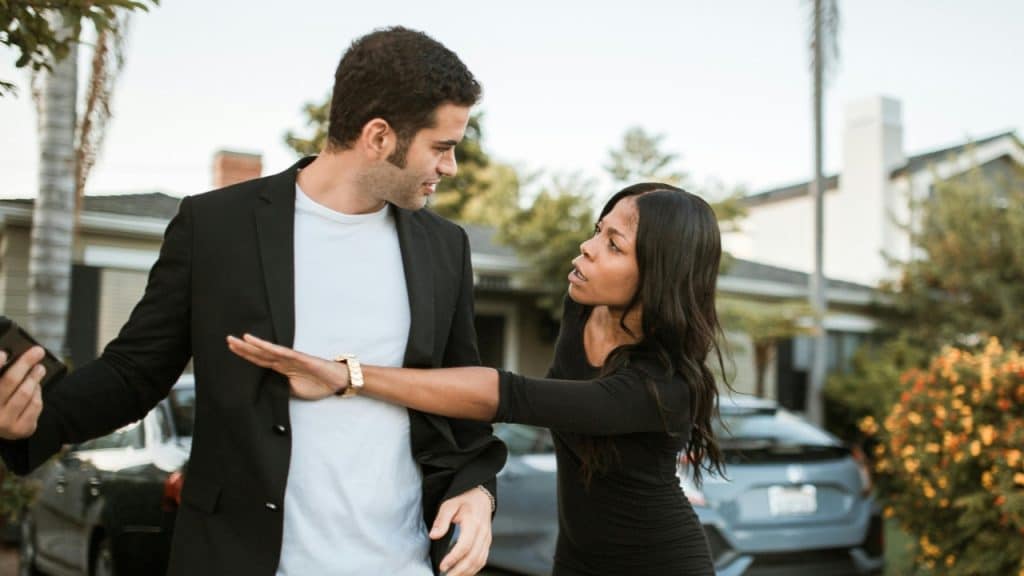 A woman reaches out to block a man holding a phone in a residential driveway.