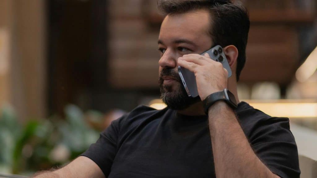 A bearded man talking on a smartphone while sitting indoors.