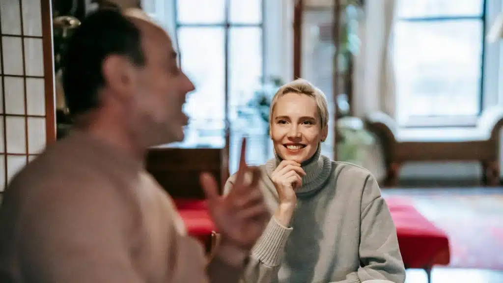 A woman in a grey turtleneck smiles while looking at a man talking in the foreground.