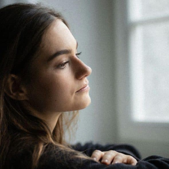 A woman with long brown hair looking out a bright window with a thoughtful expression.