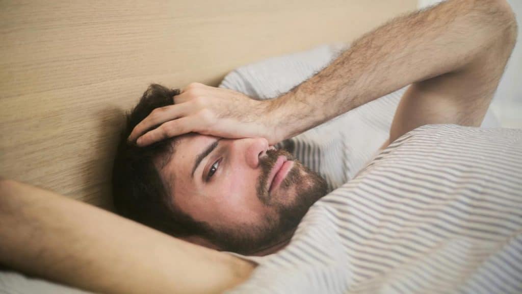 A man lying in bed with his hand on his forehead, looking unwell or tired.