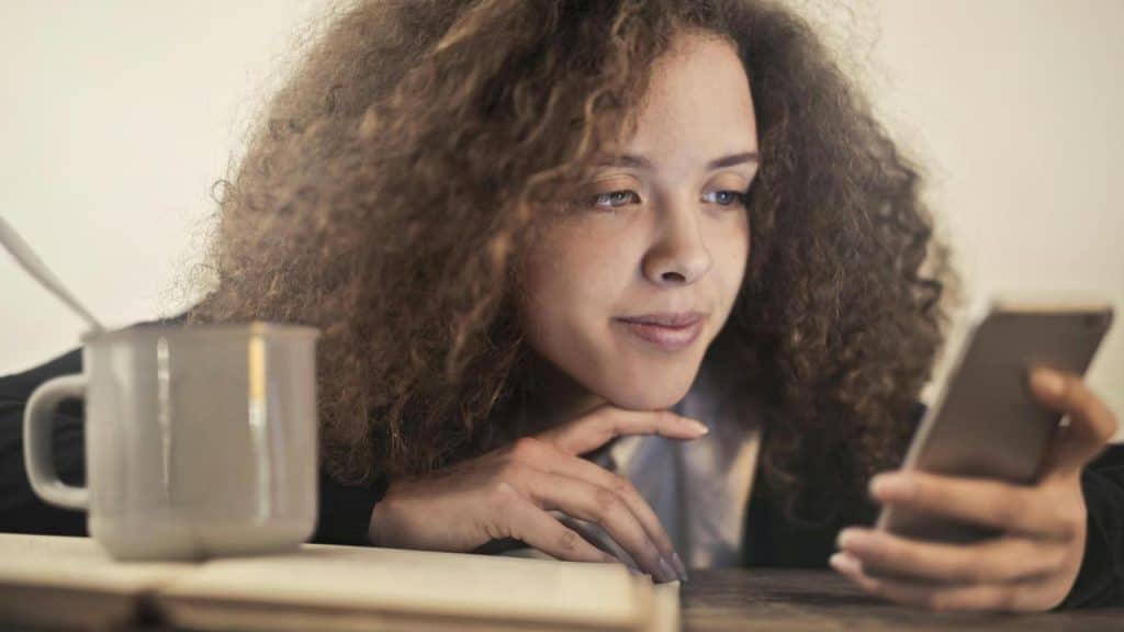 A woman with curly hair smiling while looking at her phone beside a mug.