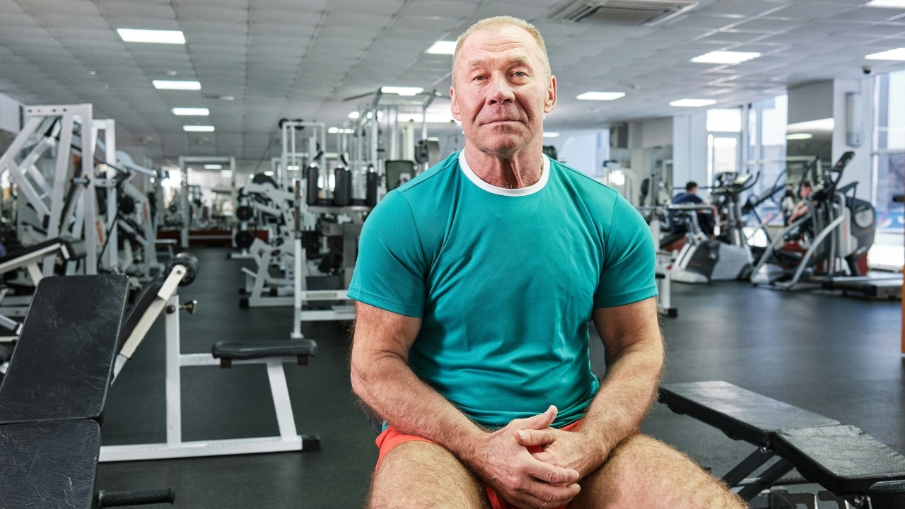 A middle-aged man in a teal shirt sits on a bench inside a large gym.