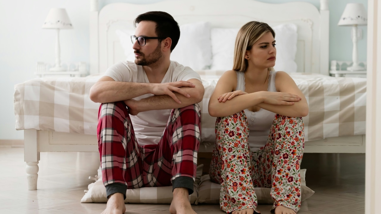 A man and woman sit on the floor against a bed, looking in opposite directions.
