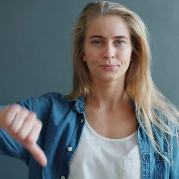 A blonde woman in a denim shirt gives a thumbs-down gesture against a grey background.