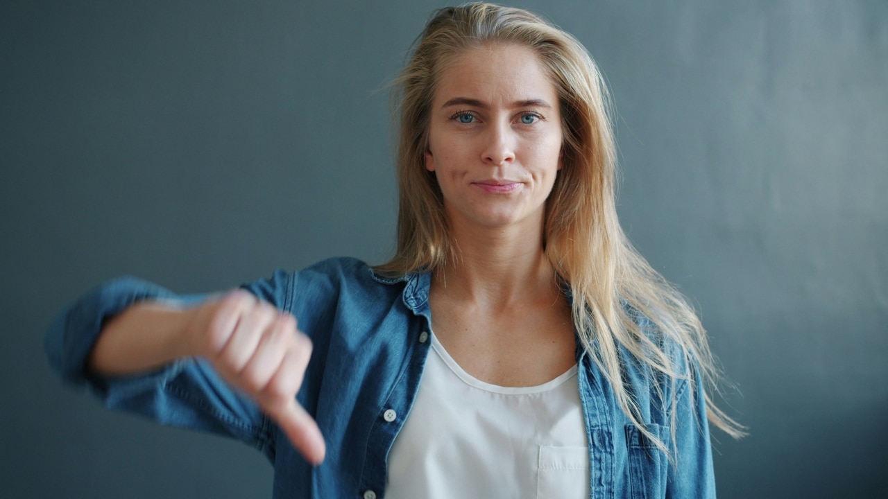 A blonde woman in a denim shirt gives a thumbs-down gesture against a grey background.