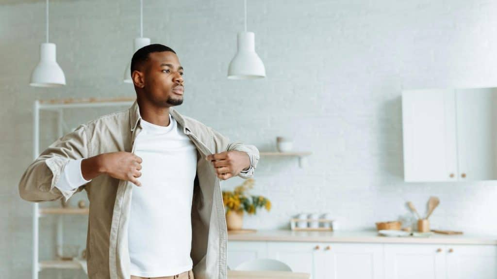 A man adjusting his shirt while standing in a bright kitchen.
