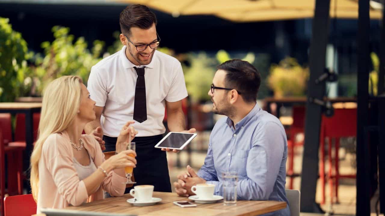 A waiter shows a tablet to a man and woman sitting at an outdoor cafe table.