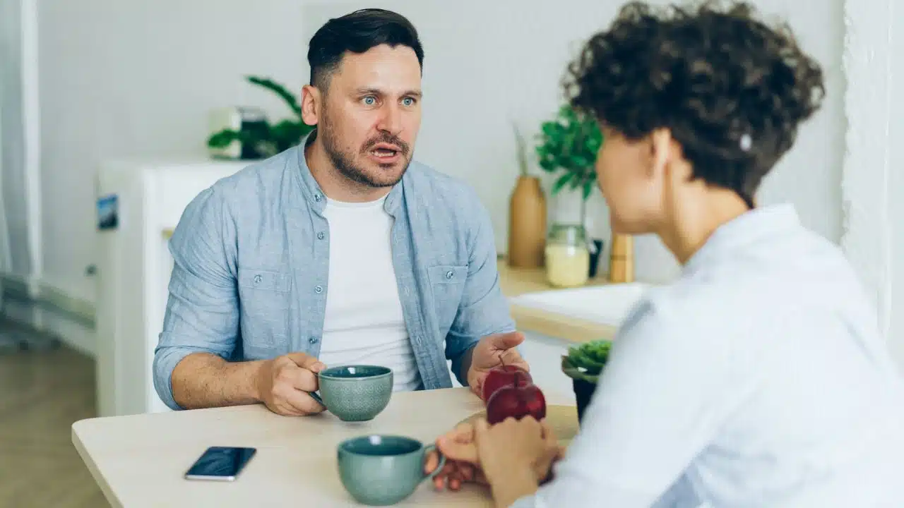 A man speaks with an open mouth while holding a mug across from a woman.