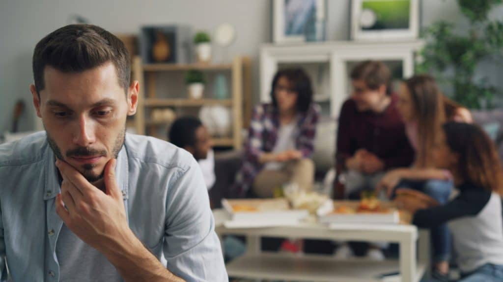 A man rests his chin on his hand while a group sits blurred in background.