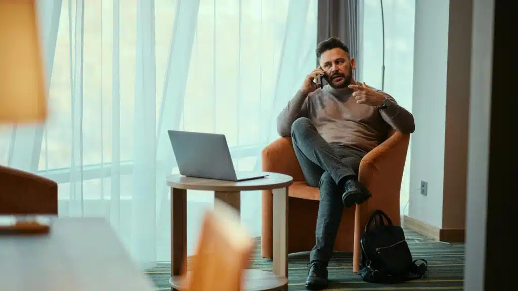 A man sitting in a chair, talking on a phone beside an open laptop.