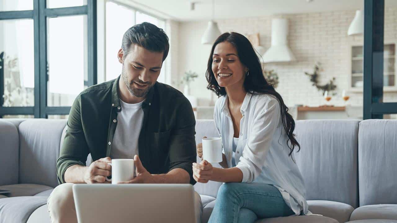 A beautiful young couple drinking coffee and looking at the laptop while sitting on the sofa at home.