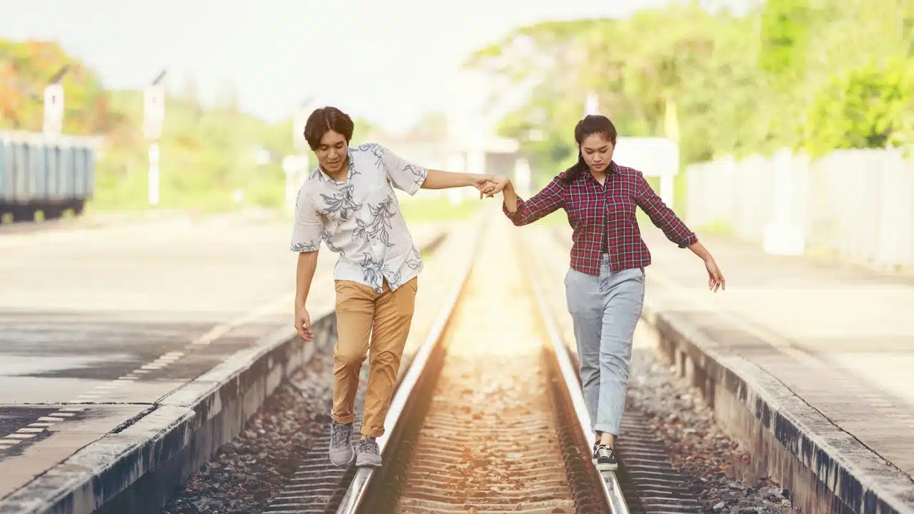 A couple walking on railway tracks while holding hands to maintain their balance at a train station.