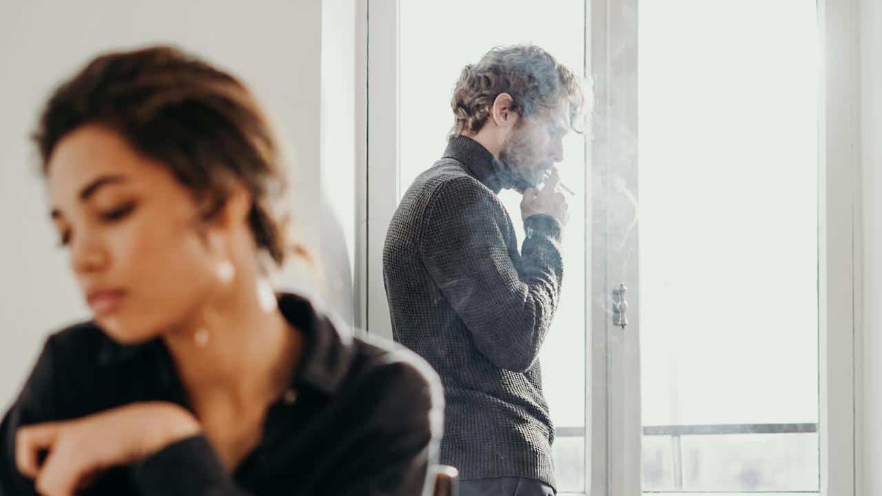 A man is standing in front of a bright window, smoking a cigarette, while a woman sits at a table in the blurred foreground
