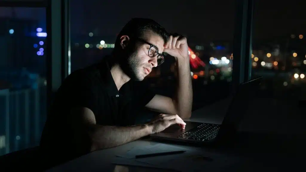 A man wearing glasses uses a laptop at night with city lights in the background.