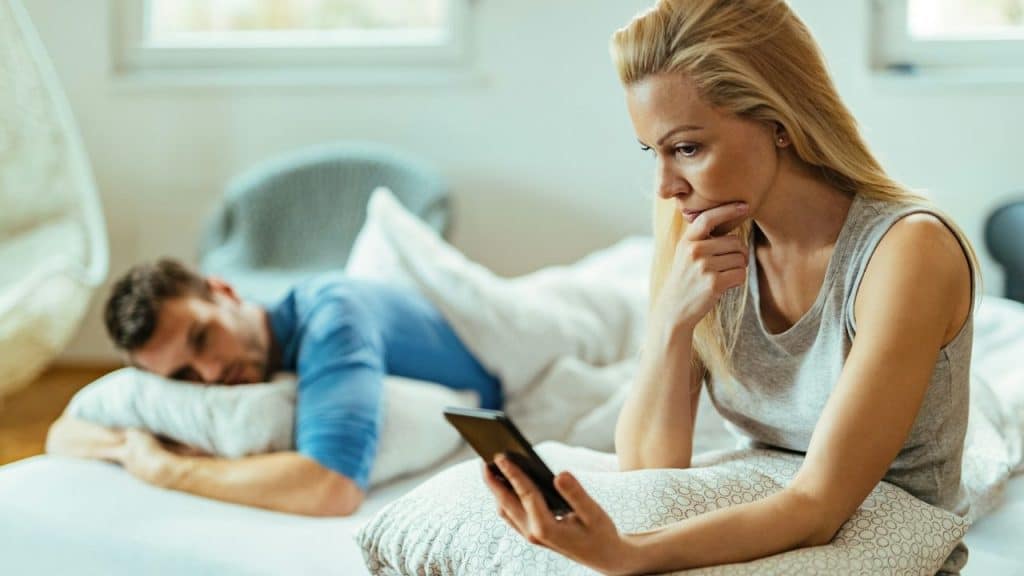 A woman looks at a smartphone while a man lies in bed in the blurred background.
