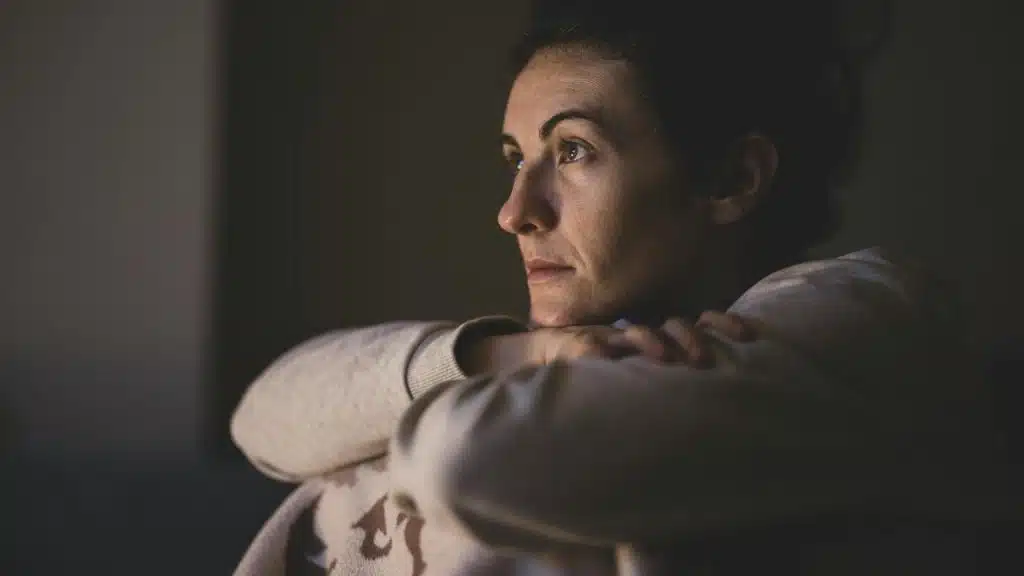 A woman sitting in dim light resting her chin on her arms and looking thoughtful.