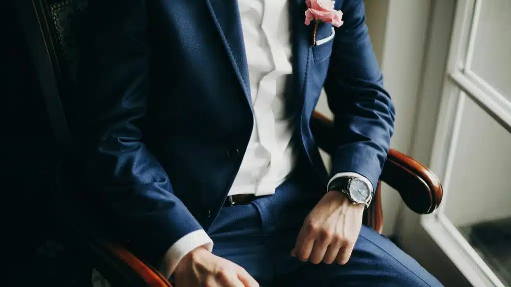 A person in a blue suit sitting by a window with a flower boutonniere.
