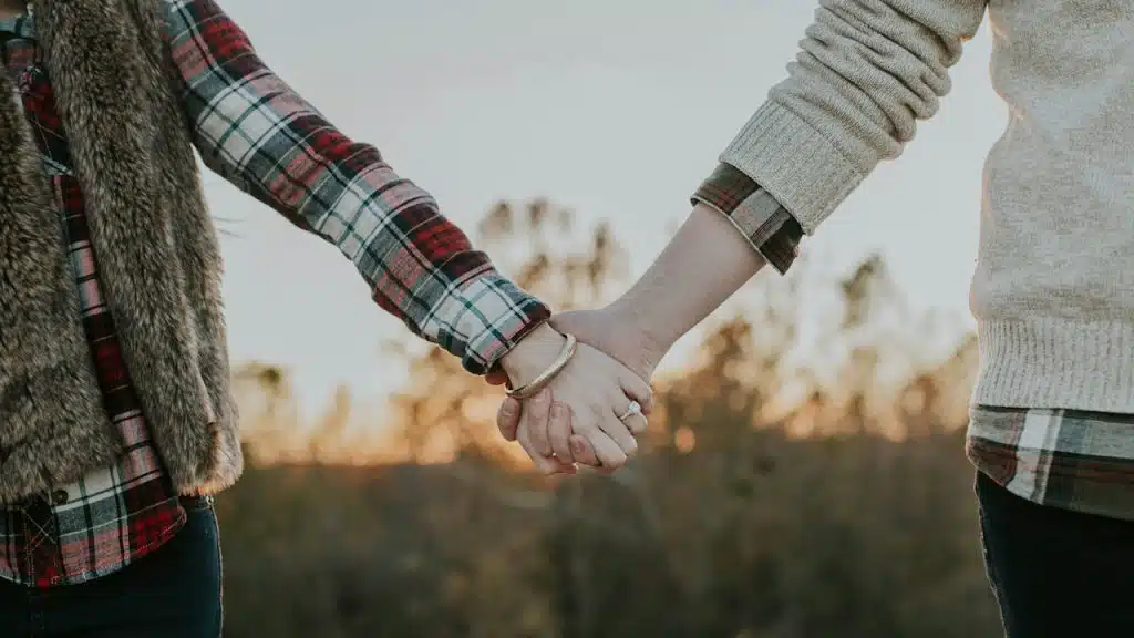 Close-up of a couple holding hands outdoors, with the sunset or golden hour light visible in the blurred background.