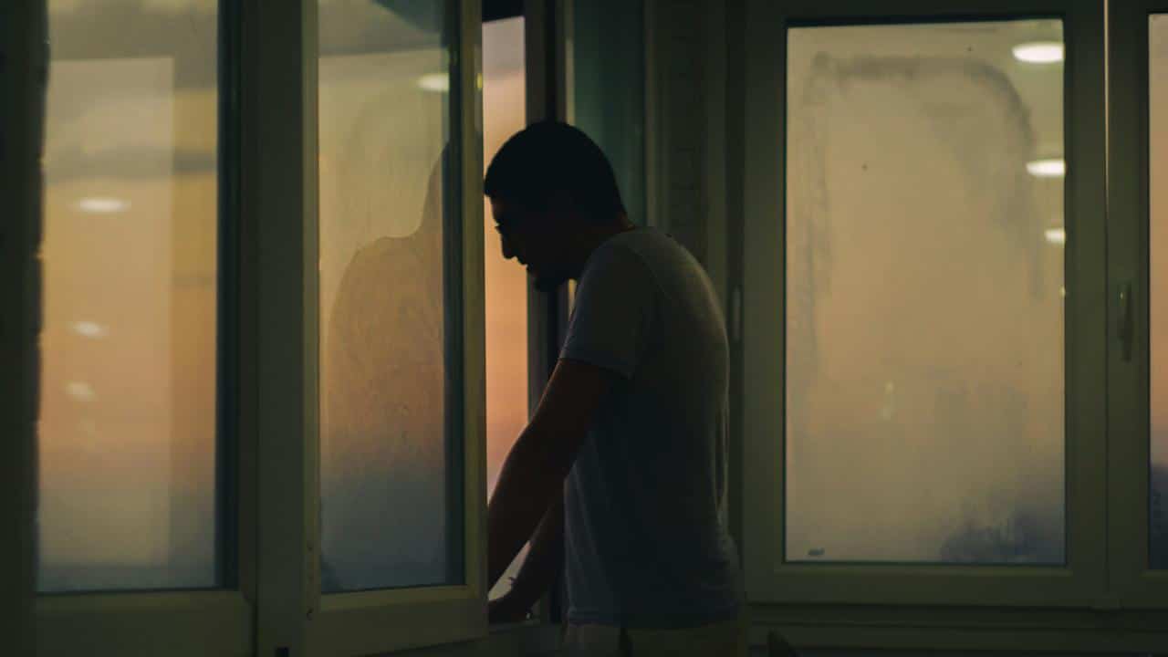 A man standing by a window in low light, looking down.