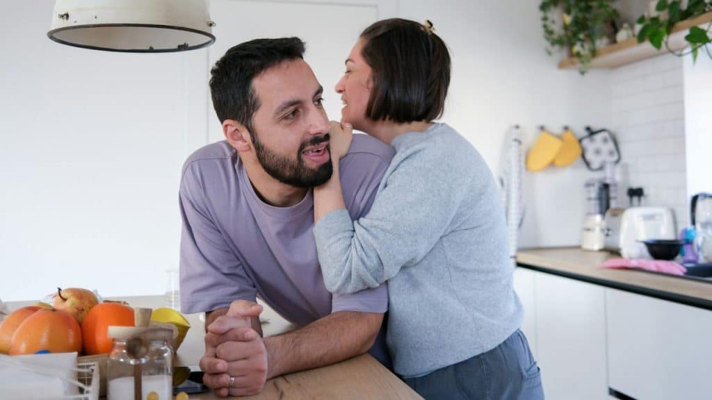 A woman leans against a man’s shoulder and whispers to him in a kitchen.