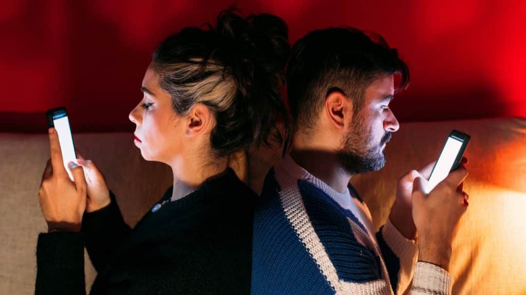 A man and woman sit back-to-back on a couch while looking at glowing smartphones.