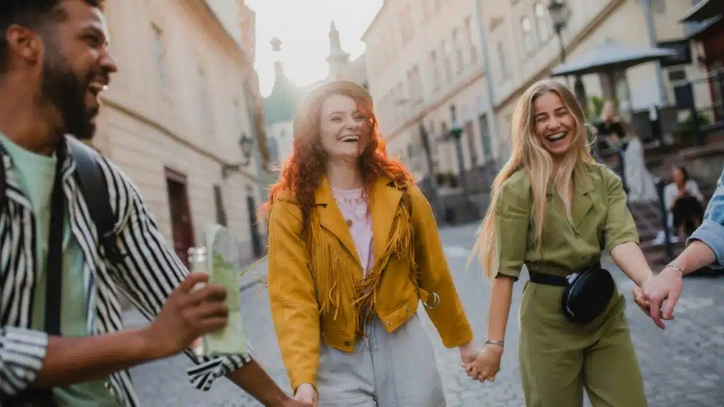 Four people hold hands and laugh while walking down a sunlit, narrow European-style street.