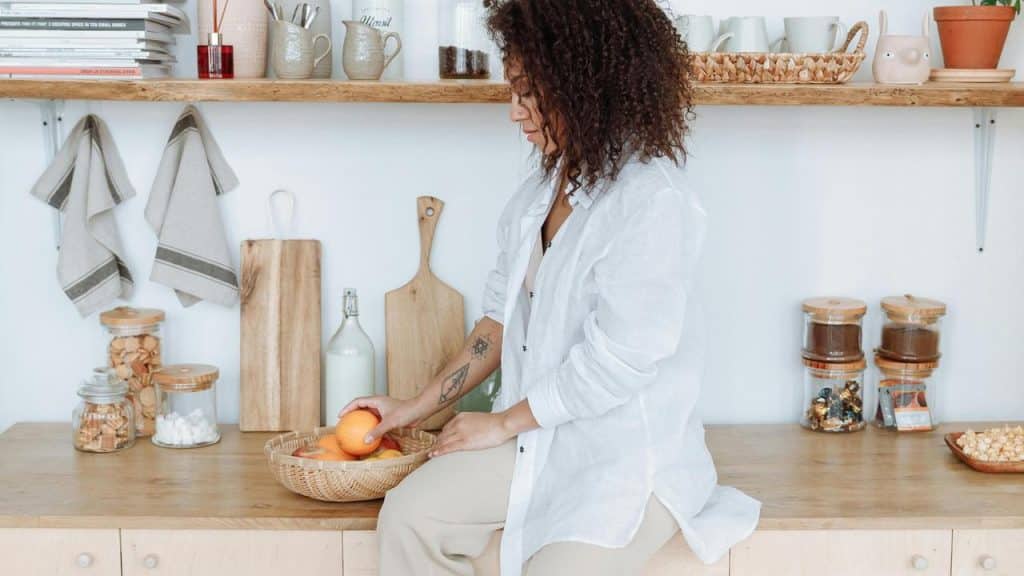 A woman sitting on a kitchen counter picking fruit from a basket.