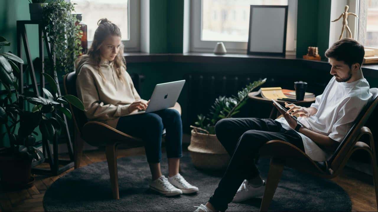A man and woman sitting in chairs, focused on their devices in a cozy room.