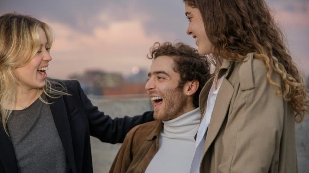 Three people stand close together outdoors, laughing and smiling under a soft, evening sky.
