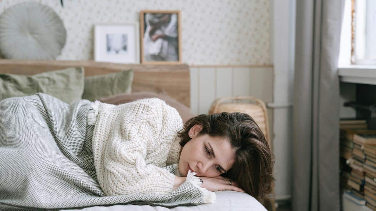 A woman lies on a bed wrapped in a blanket, looking sad.