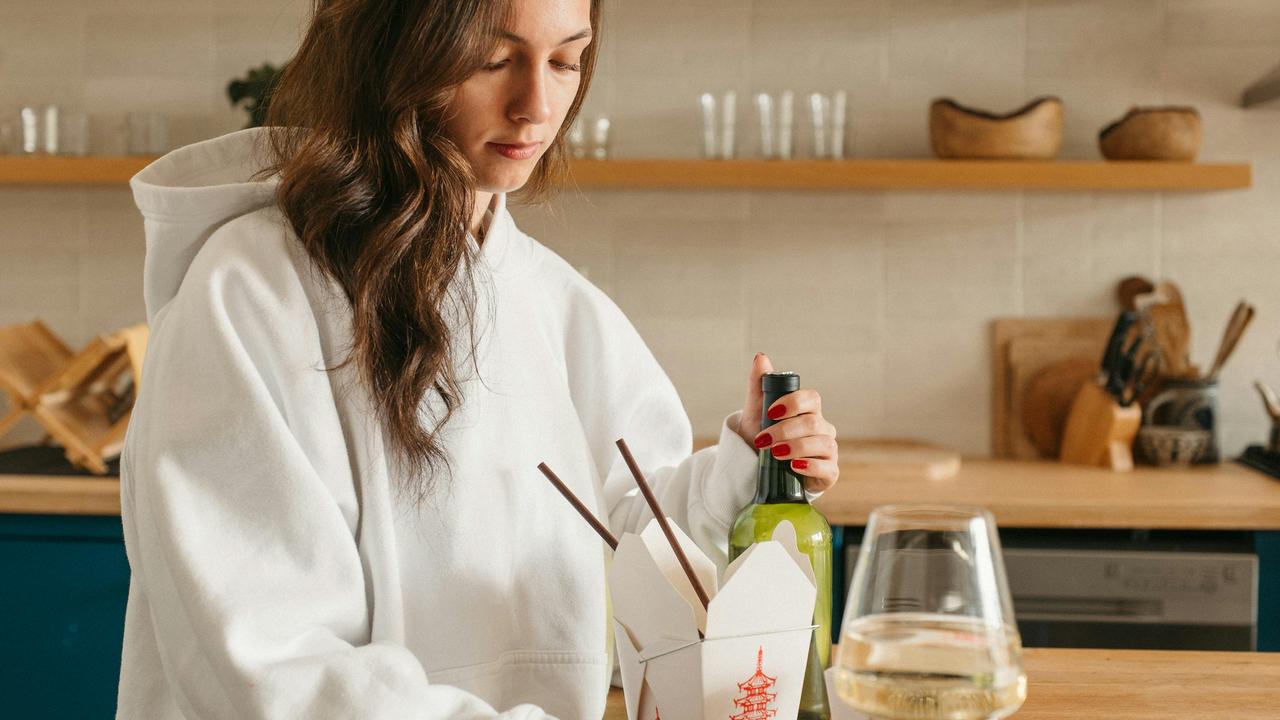 A woman opening a bottle beside a takeout container and wine glass.