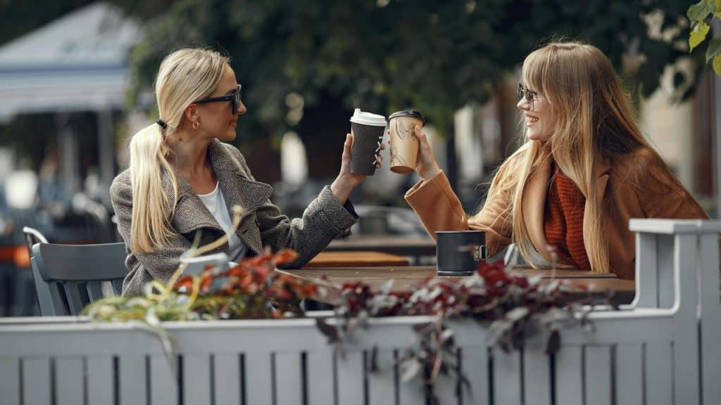 A woman sitting at an outdoor café, smiling and clinking coffee cups with another woman.