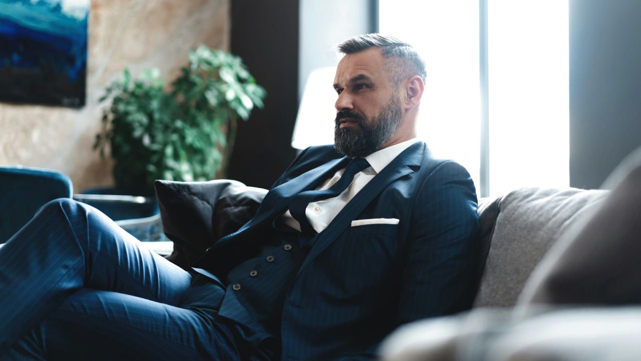 A man in a blue three-piece suit sits on a couch and looks toward a window.
