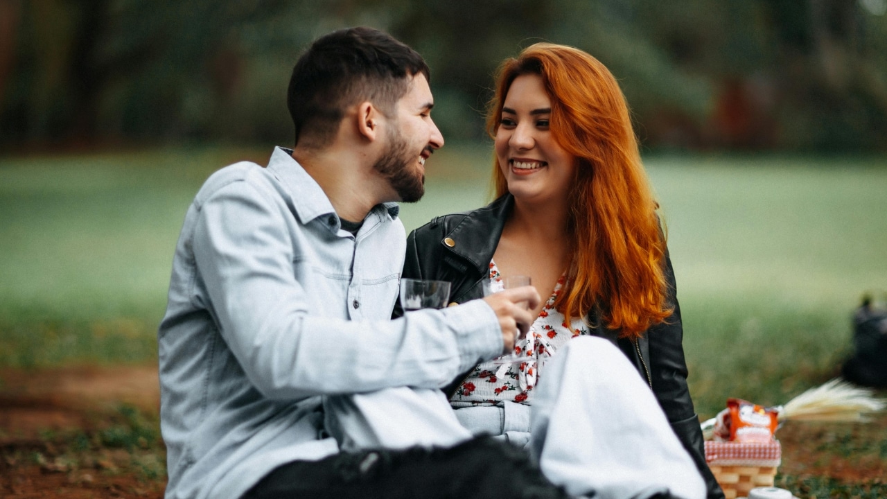 A man and a woman with red hair sit on the grass laughing during a picnic.